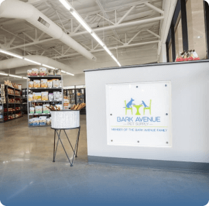 Modern pet store interior with a gray floor, high ceilings, and bright lighting. A sign reading "Bark Avenue" is on a wall near shelves of pet supplies.
